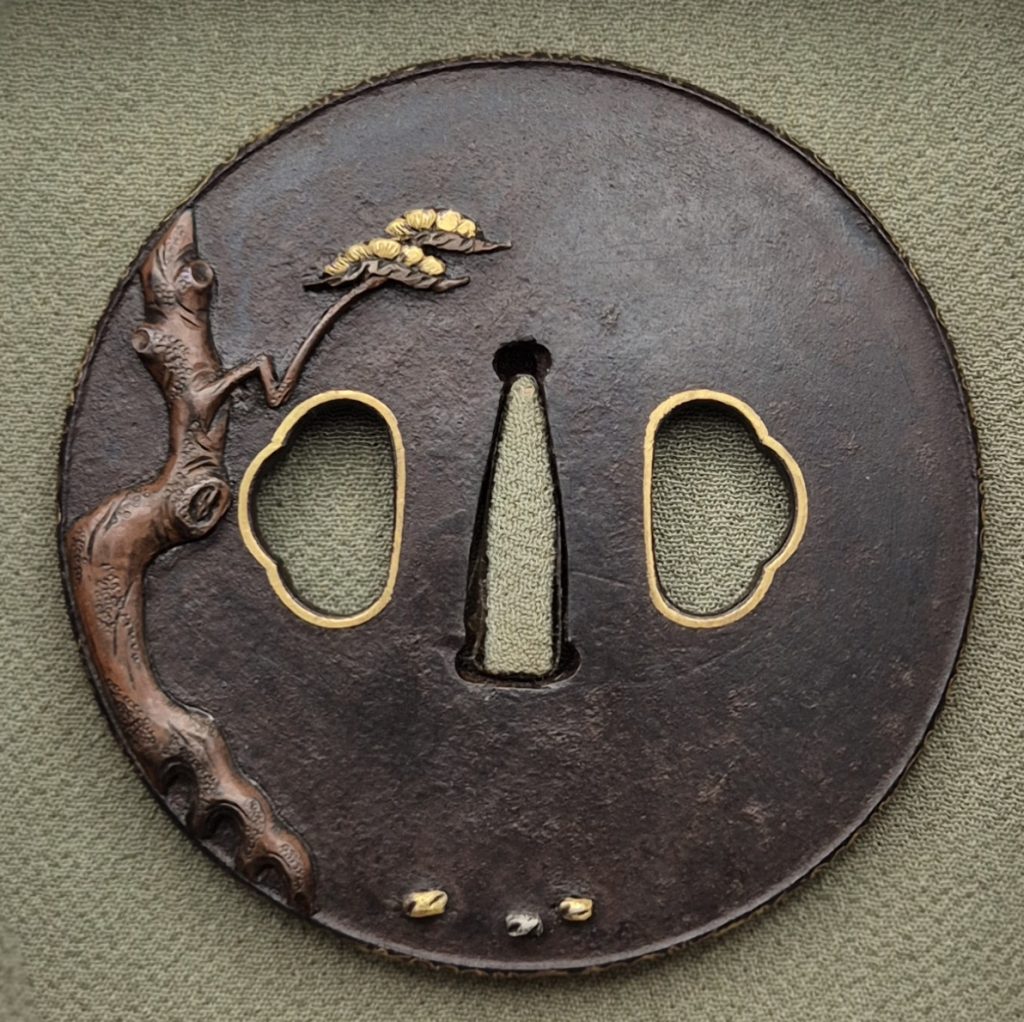 The back of the iron tsuba: a reddish pine trunk rises along the left edge to a single gold cluster of needles, with three small inlays below the opening.
