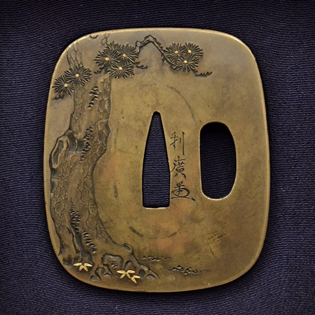 The back of the brass tsuba: an old pine rises along the left edge, small gold grasses at its foot, and the signature Toshihiro with kao beside the central opening.