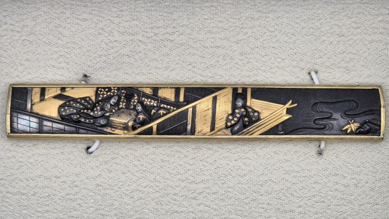 Shakudo kozuka with a scene from Genji, resting in its box: ladies in patterned robes around a go board, a gold veranda across the middle, dark water with a small spray of gold bamboo on a stone at the right.