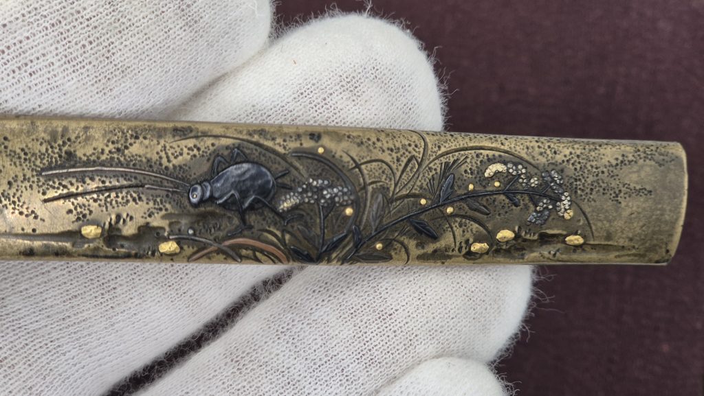 Detail of the cricket in shakudo with long silver antennae, the grasses engraved and inlaid in copper and shakudo, gold pebbles along the lower edge.