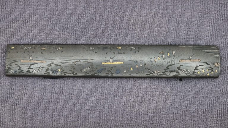 The Hosono Masamori kozuka: a river landscape in flat inlay on grey shibuichi, tiny figures in straw hats moving along the banks, trees, and short bridges of gold and copper across the water.