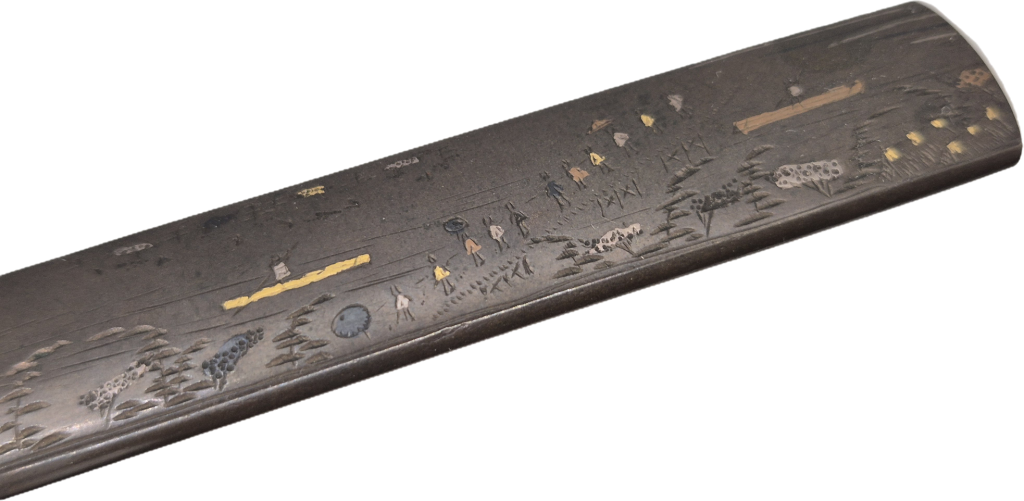 Detail of the kozuka at an angle: a file of travellers in triangular hats, a gold bridge and a round dark inlay, trees in silver and shakudo.