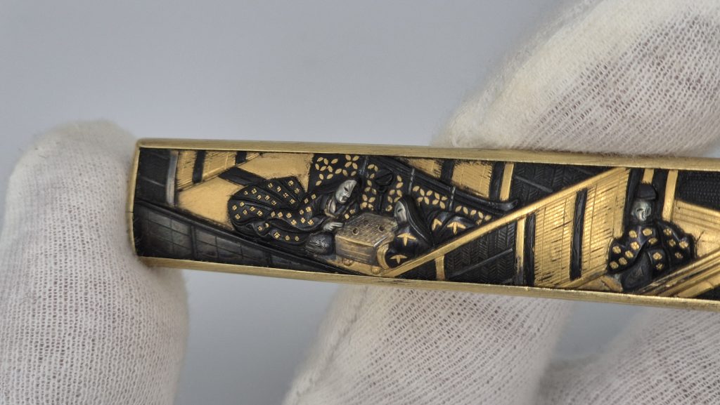 The kozuka held in a gloved hand, the palace scene in raking light.
