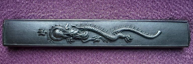 Kaga Goto kozuka in shakudo with a crawling dragon in high relief on a nanako ground, the jewel at the head end.
