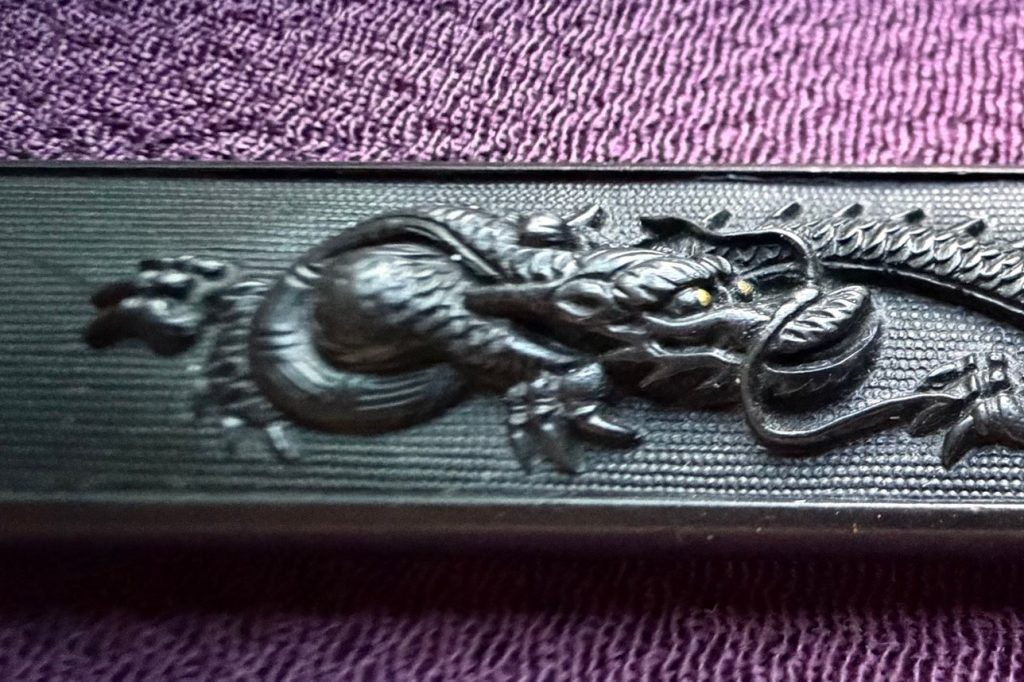 The kozuka seen at a low angle, showing the depth of the relief against the granulated ground.