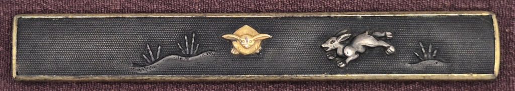 The rabbit kozuka: on a dark nanako ground a gold hare crouches facing the viewer with its ears spread, a silver hare leaps to the right, tufts of grass at both ends.