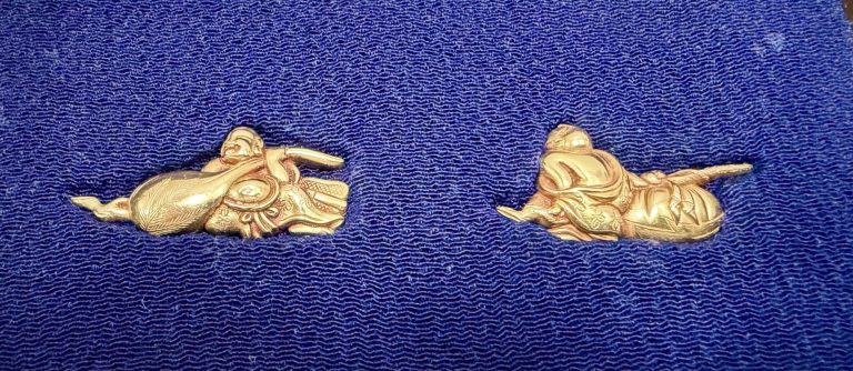 The pair of gold menuki on dark blue silk, each a stout reclining figure of Hotei with his great bag.