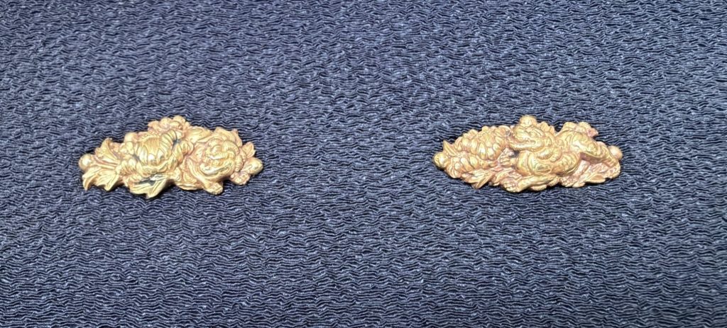 The pair of botan shishi menuki in gold on dark blue silk, each a cluster of layered peony blossoms with a shishi half hidden among them.