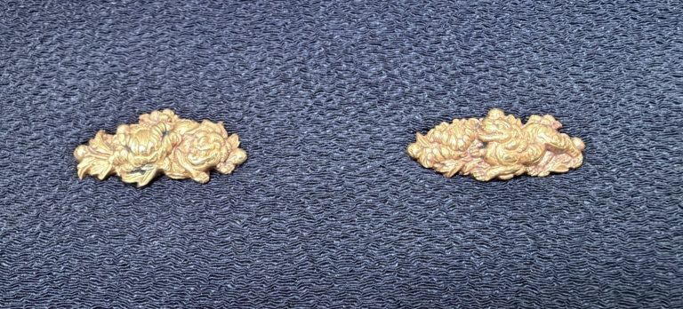 The pair of botan shishi menuki in gold on dark blue silk, each a cluster of layered peony blossoms with a shishi half hidden among them.