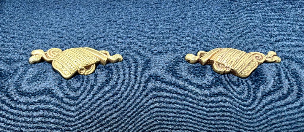 The pair of osode menuki in gold on blue silk, each a large shoulder guard of armor with laced rows of plates and a cord curling away at either end.