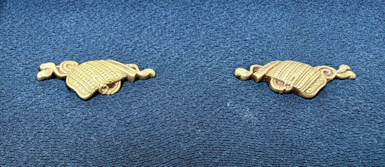 The pair of osode menuki in gold on blue silk, each a large shoulder guard of armor with laced rows of plates and a cord curling away at either end.