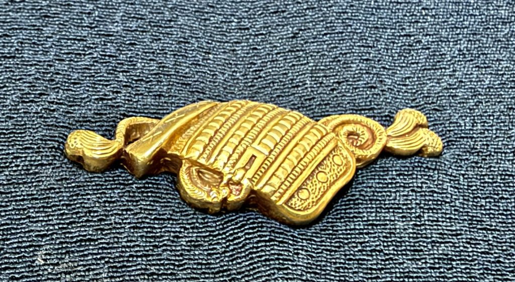 The other menuki in warm light: the shoulder guard lies slightly folded, with a border of small rings along its lower edge.