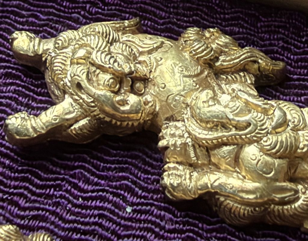 Detail of one menuki: a shishi with round staring eyes faces out at the left, while a second shishi presses its curled mane against the first one's spotted flank.
