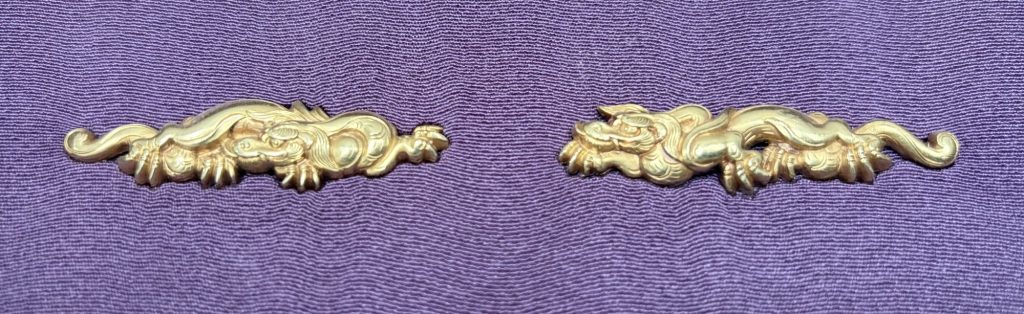 The pair of Mito menuki in gold on purple silk: two rain dragons laid out as mirror images, the tails curling outward at either end.