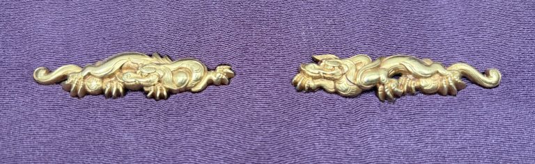 The pair of Mito menuki in gold on purple silk: two rain dragons laid out as mirror images, the tails curling outward at either end.