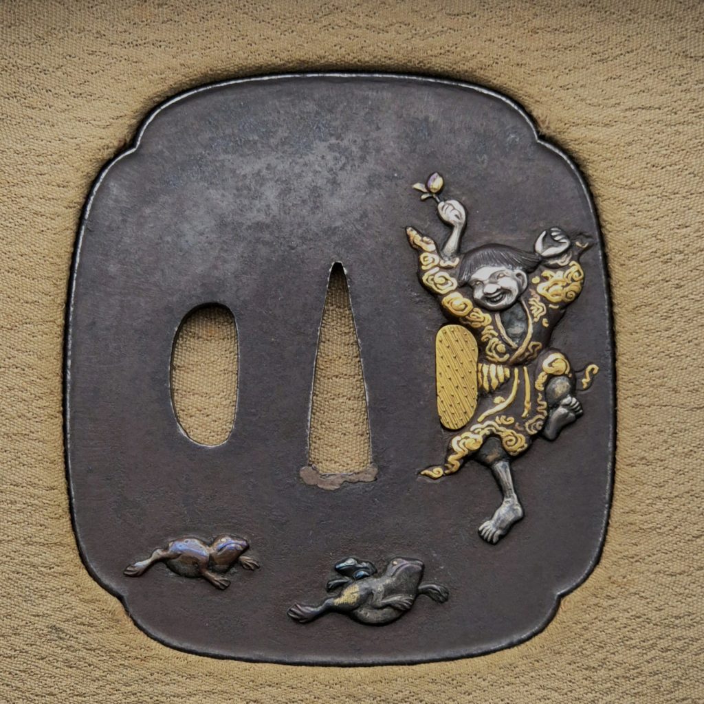 The front of the iron Gama Sennin tsuba: the laughing immortal dances on one leg at the right, a bell raised in his hand, two toads crossing the lower edge.