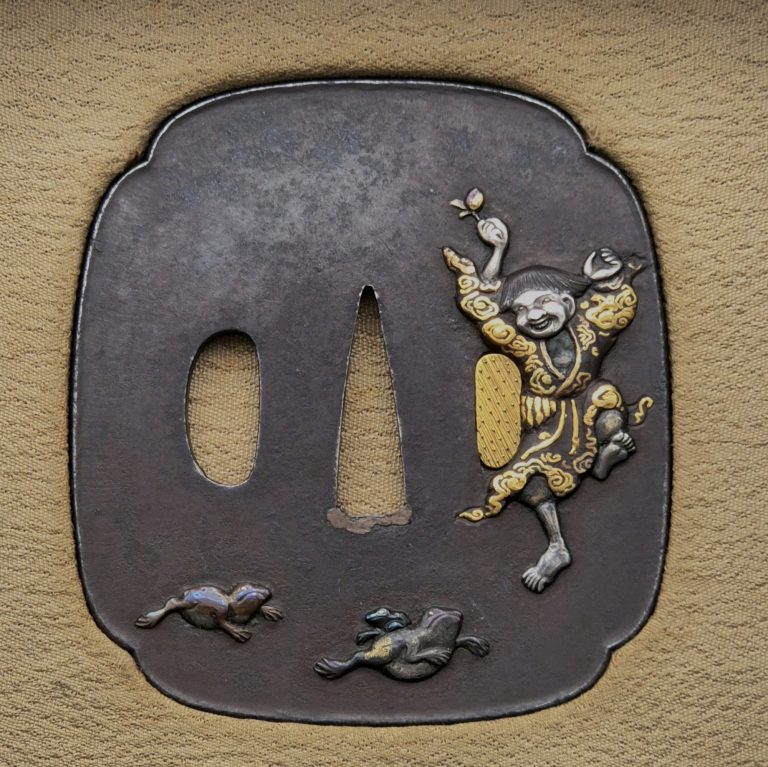 The front of the iron Gama Sennin tsuba: the laughing immortal dances on one leg at the right, a bell raised in his hand, two toads crossing the lower edge.