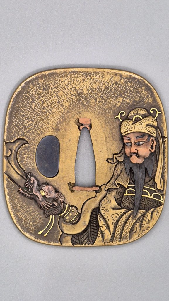 The front of the Guan Yu tsuba by Tsuneshige: the general at the right edge with copper face, black beard and gold headdress, the blade of his halberd rising from a dragon's head at the left.
