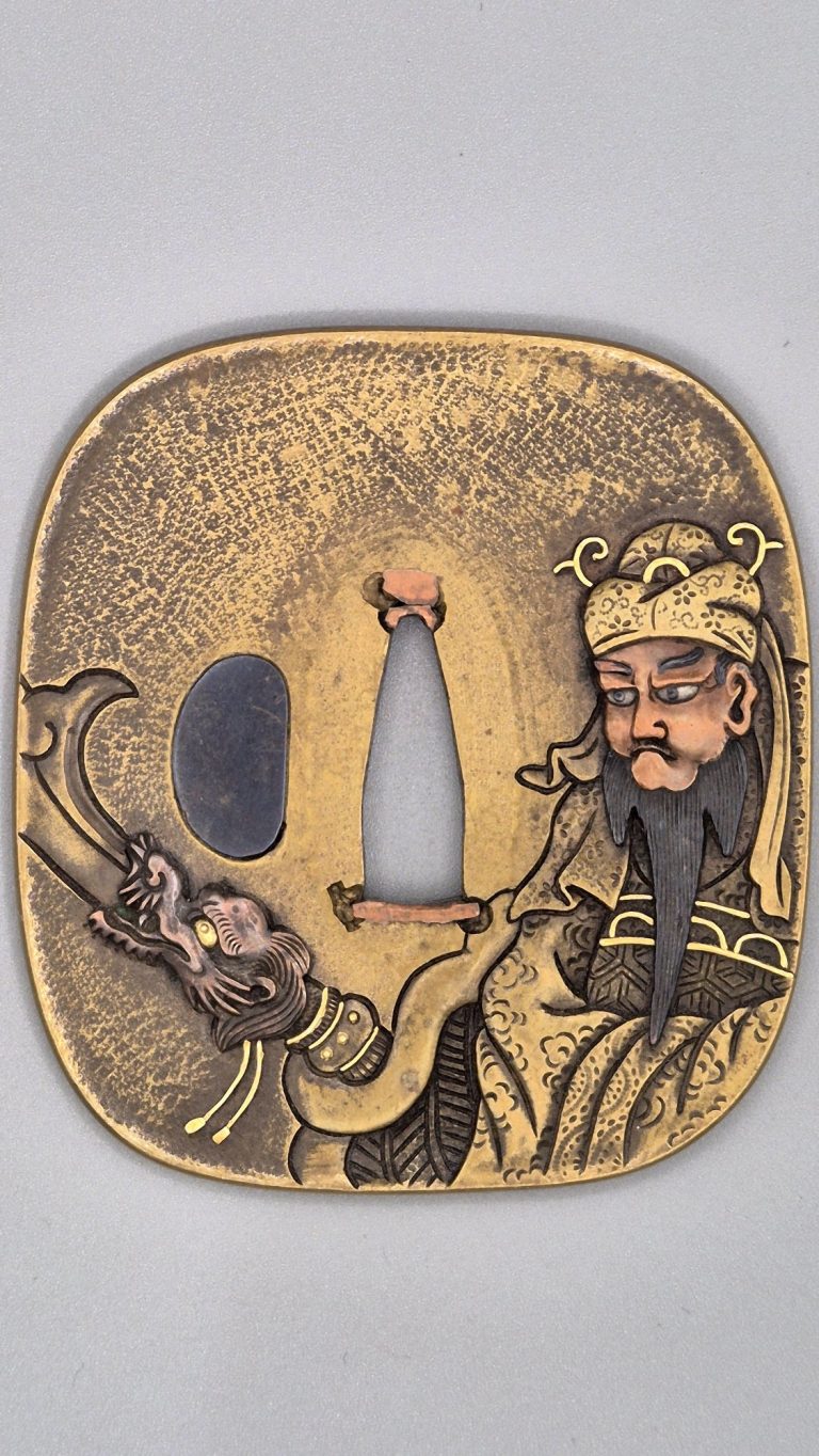 The front of the Guan Yu tsuba by Tsuneshige: the general at the right edge with copper face, black beard and gold headdress, the blade of his halberd rising from a dragon's head at the left.