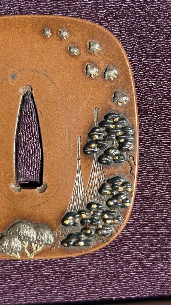 Detail of the front: the plovers in flight, the pine with gold needles and the fine engraved lines of the nets.