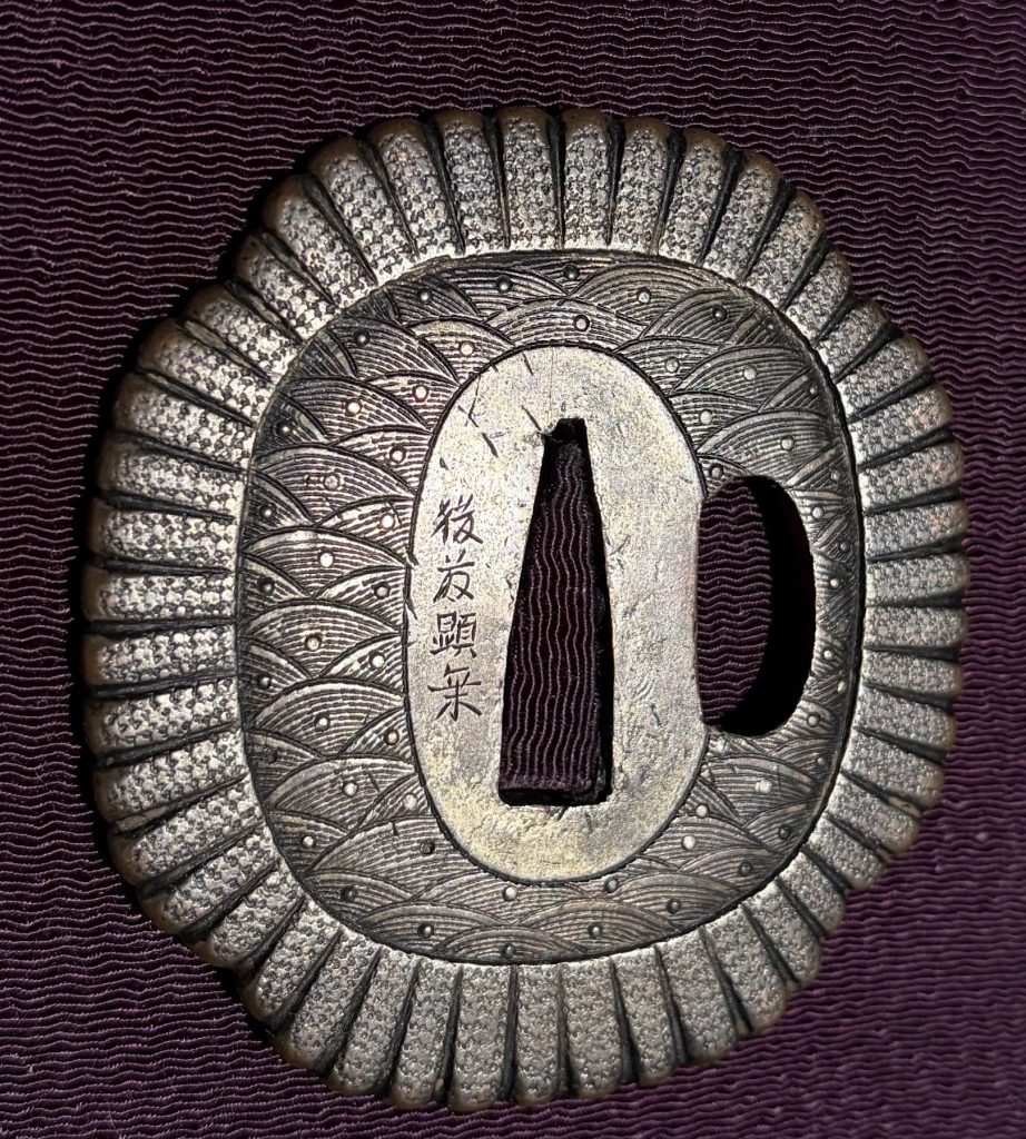 The Ko-Kinko tsuba: rows of seigaiha waves inside a rim of ribbed chrysanthemum petals, a signature beside the tang opening.