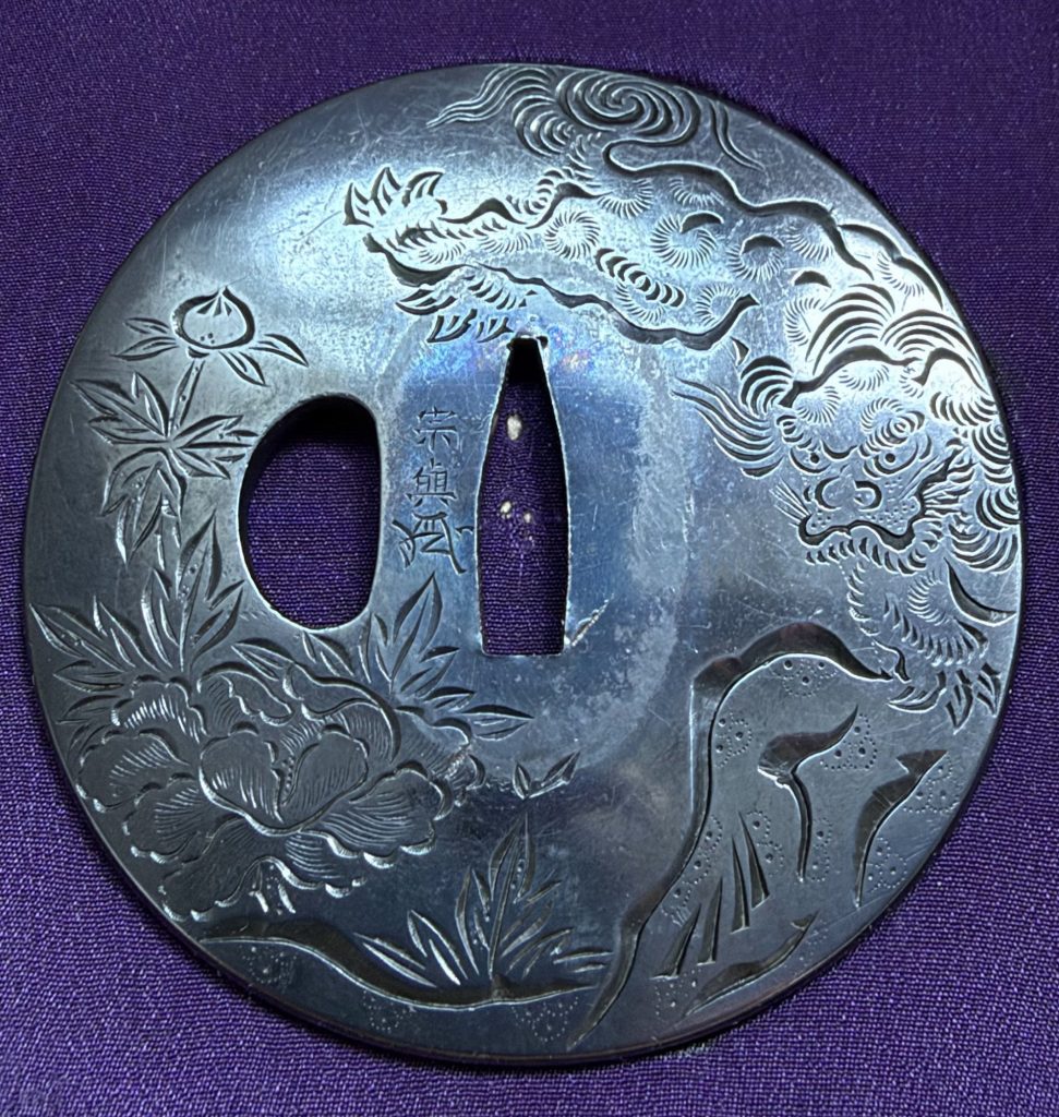 The front of the Yokoya Soyo tsuba: a shishi on the right, a flowering peony on the left, all cut in katakiri-bori on a polished plate.