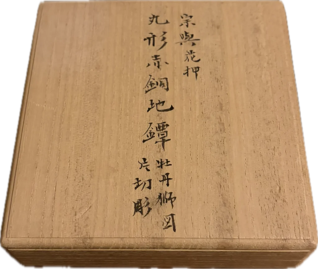 The lid of the storage box, inscribed in ink: Soyo with kao, a round tsuba on a shakudo ground, peony and shishi, katakiri-bori.