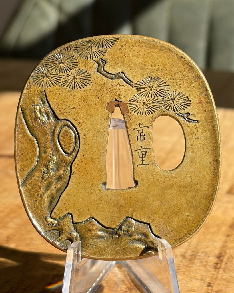 Reverse of the tsuba: pine branches, an old trunk with a knot hole, low hills and the signature 常重 beside the blade opening.