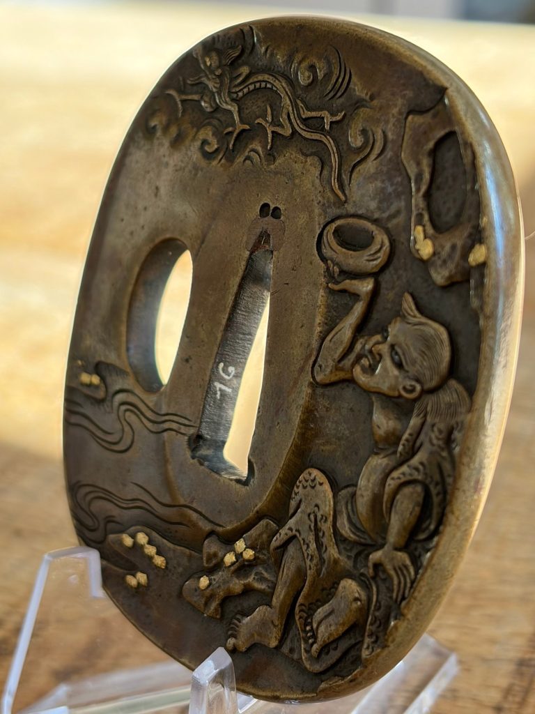 The tsuba at an angle, showing the thick rim, gold dots on the rocks and the number painted inside the blade opening.