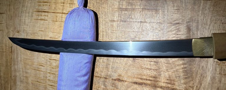 Hira-zukuri wakizashi blade on violet silk across a wooden table, an undulating notare hamon above a gold-toned habaki.